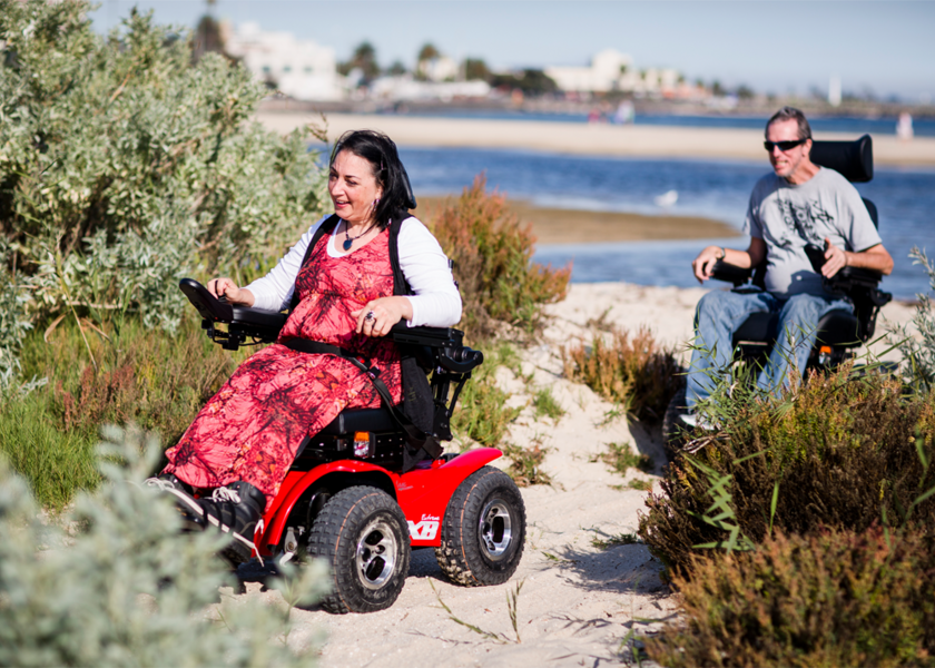 Zwei Personen im Rollstuhl fahren am Strand