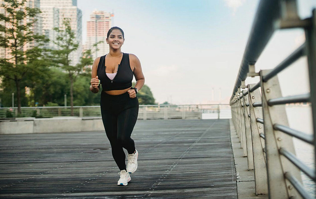 Beitragsfoto zu "Knieschmerzen beim Laufen" - Abbildung einer Joggerin, die draußen in einer städtischen Umgebung am Wasser joggen geht.