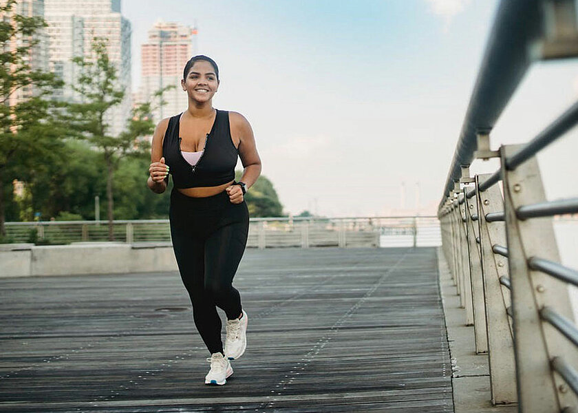 Beitragsfoto zu "Knieschmerzen beim Laufen" - Abbildung einer Joggerin, die draußen in einer städtischen Umgebung am Wasser joggen geht. 